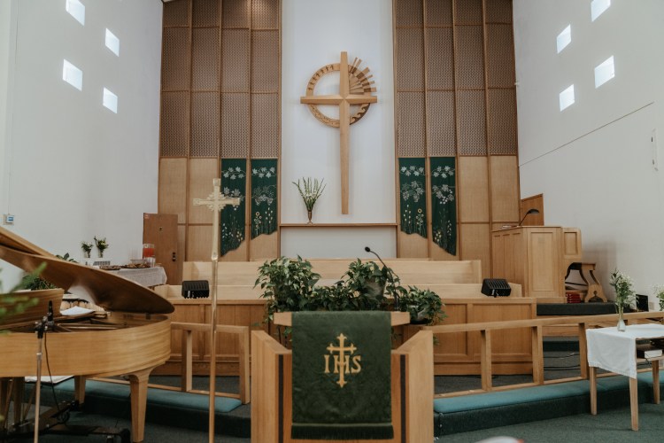 Wide view of a light wood altar covered with dark green altar cloth. A Christian cross is on the wall in the background.